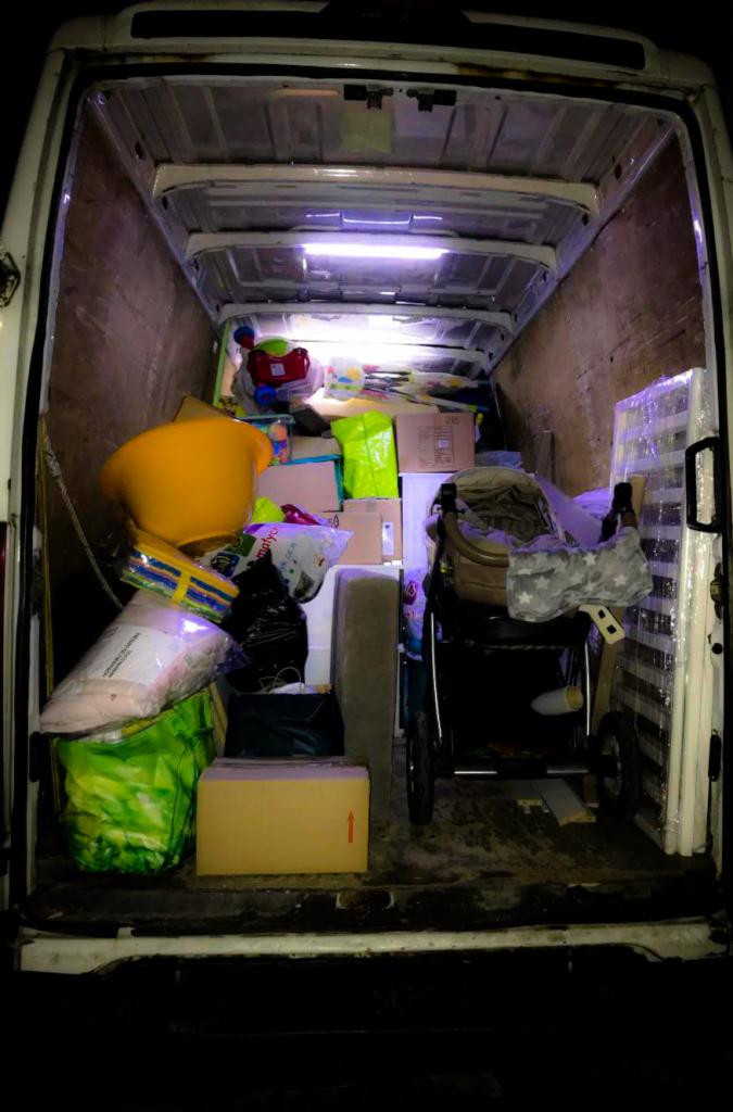 Wnętrze białego busa załadowane kartonami, meblami i wózkiem dziecięcym, oświetlone górnym światłem, widok z tyłu pojazdu.