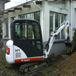 Minikoparka Bobcat 321 z łyżką, częściowo zanurzona w ziemi, na tle budynku z dużymi oknami, w trakcie prac ziemnych.