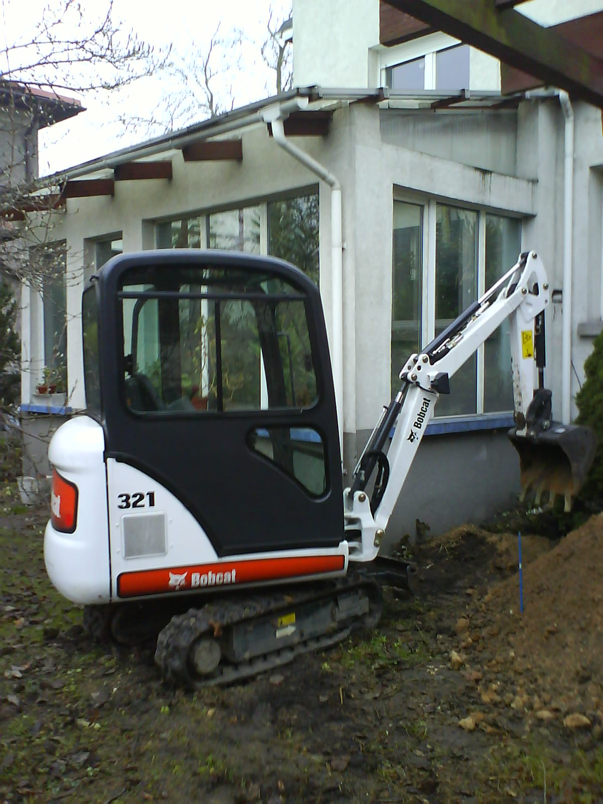 Minikoparka Bobcat 321 z łyżką, częściowo zanurzona w ziemi, na tle budynku z dużymi oknami, w trakcie prac ziemnych.