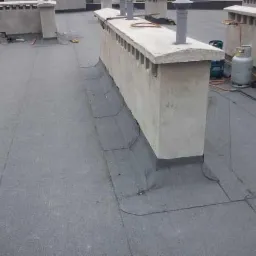 Szary dach pokryty papą z widocznymi kominami, narzędziami dekarskimi i butlą gazową w tle.