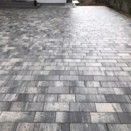 Ułożona szara kostka w kształcie prostokątów tworząca dużą powierzchnię, widoczne schody wejściowe do budynku w tle.