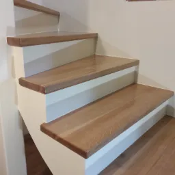 Drewniane schody z białymi podstopnicami, widoczne fragmenty ceglanej ściany w tle.