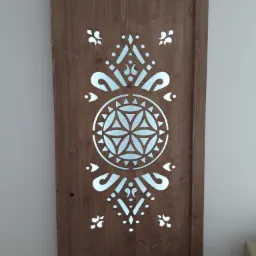 Drewniane drzwi z wyciętym dekoracyjnym wzorem geometrycznym w kolorze srebrnym, widoczne słoje drewna.