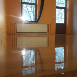 Błyszcząca, świeżo polakierowana podłoga drewniana odbija okna i kaloryfer w pomarańczowym pokoju; widoczny czarny wąż wentylacyjny przy oknie.