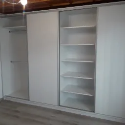 Wnętrze szafy wnękowej z przesuwnymi drzwiami, widoczne półki i drążki na ubrania, drewniany sufit z lampami w tle.