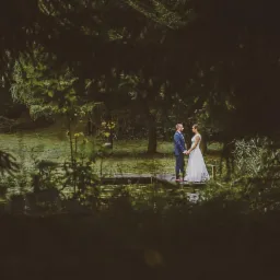 Para młoda trzymająca się za ręce na drewnianym pomoście nad stawem, otoczona zielenią drzew i krzewów, widok przez gałęzie na pierwszym planie.