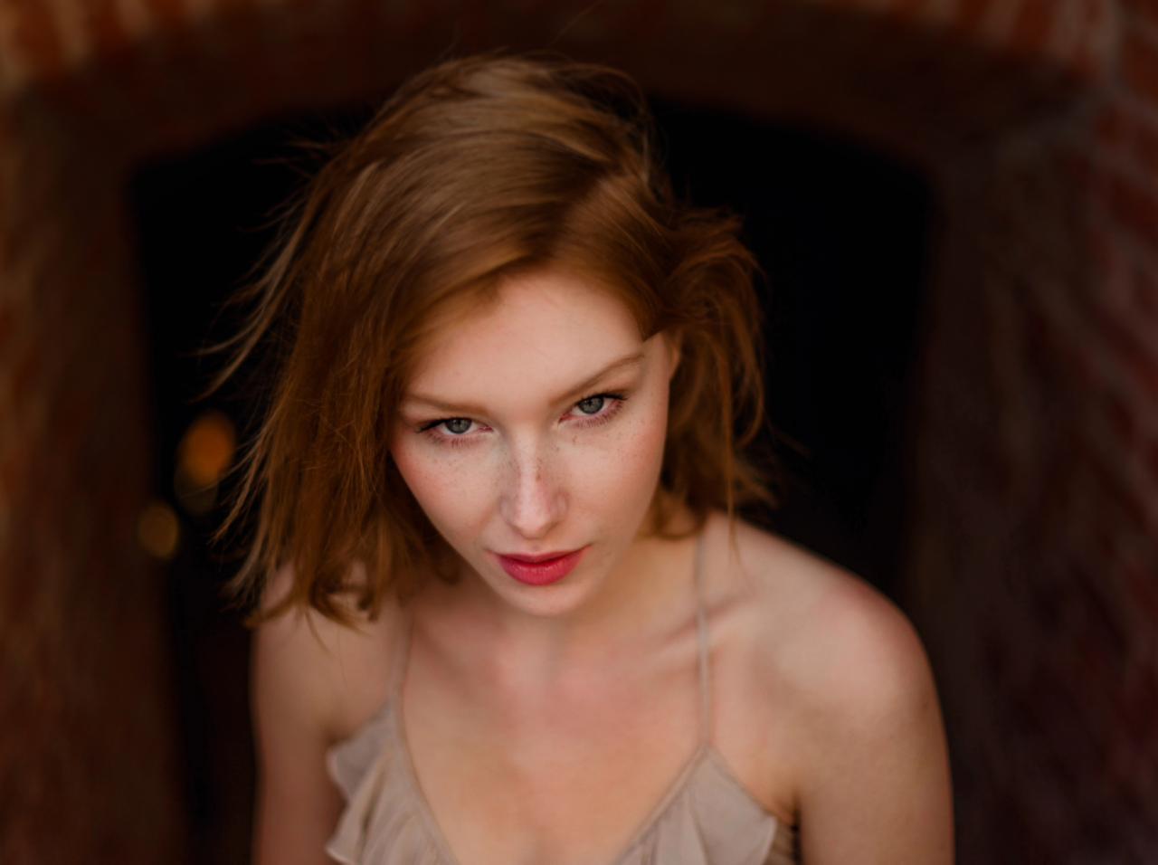 Portret młodej kobiety z rudymi włosami i piegami, spoglądającej w dół, na tle ceglanego łuku.