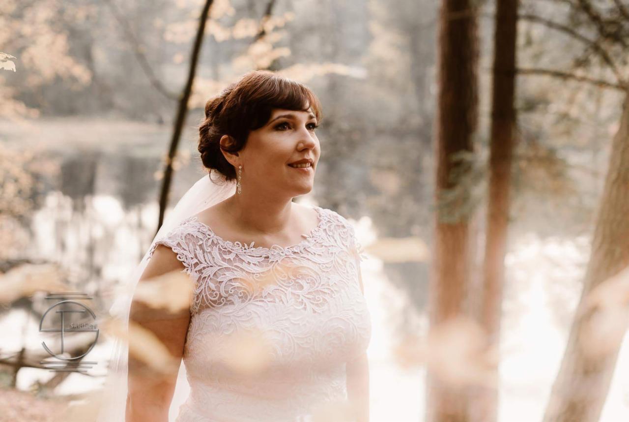 Panna młoda w koronkowej sukni i welonie, z kolczykami, spogląda w dal na tle jeziora i drzew w lesie, jesienna sceneria.