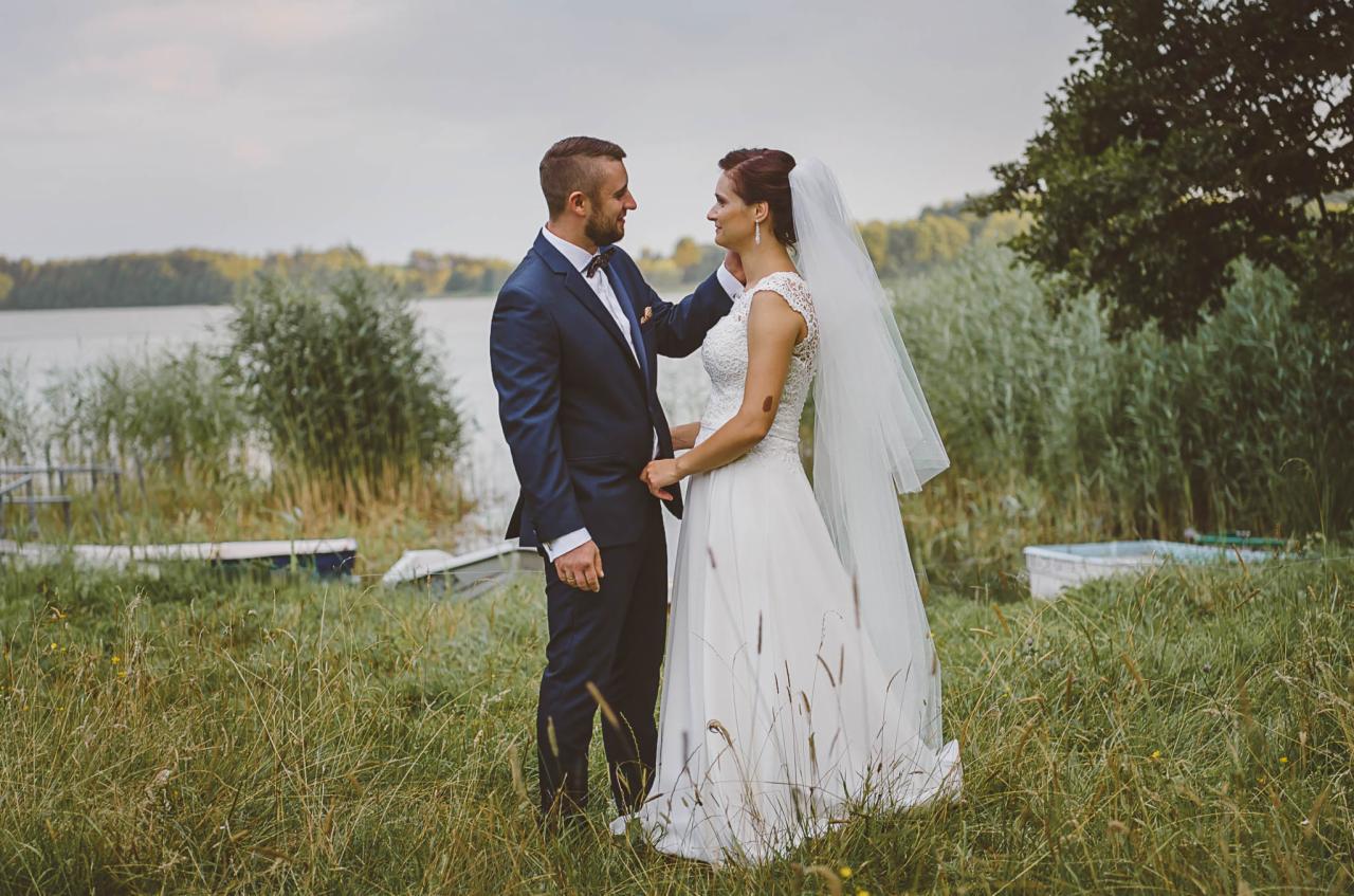Para młoda w Działdowie pozuje na tle jeziora, panna młoda w koronkowej sukni i welonie, pan młody w granatowym garniturze z muszką.