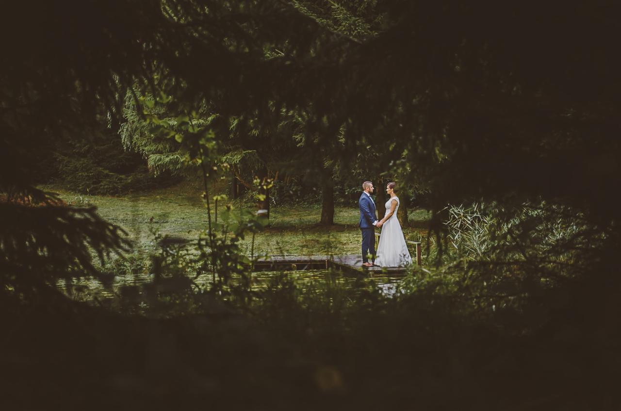 Para młoda trzymająca się za ręce na drewnianym pomoście nad stawem, otoczona zielenią drzew i krzewów, widok przez gałęzie na pierwszym planie.