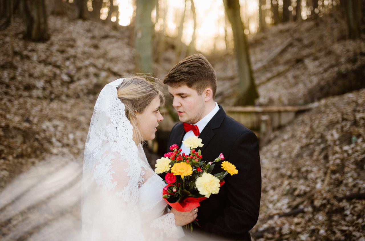 Para młoda w Działdowie, w jesiennej scenerii leśnej, spogląda na siebie trzymając bukiet z żółtych i czerwonych kwiatów. Pan młody w czarnym garniturze z czerwoną muchą, panna młoda w białej sukni...