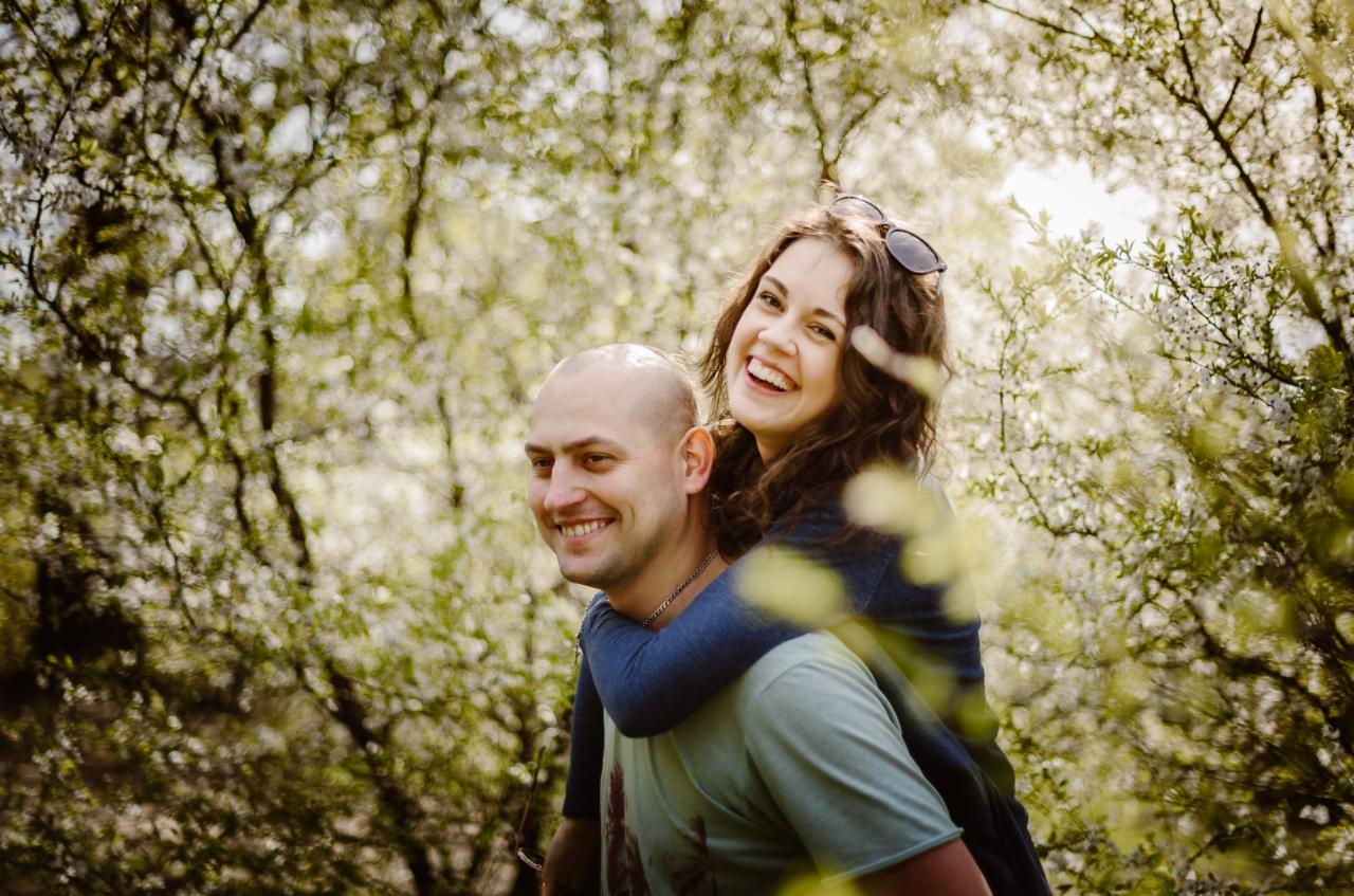 Uśmiechnięta kobieta na barana u mężczyzny w kwitnącym sadzie, okulary na głowie, radosna atmosfera.