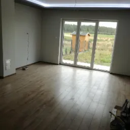 Pusty pokój po remoncie z panelami podłogowymi, widoczne okno tarasowe z widokiem na ogród i narzędzia malarskie w rogu.