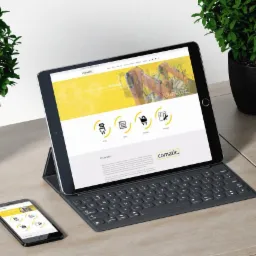 Tablet i smartfon wyświetlające stronę internetową Comatic z żółto-białym designem, obok słuchawki Bang & Olufsen i doniczki z roślinami na drewnianym biurku.