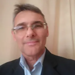 Portret mężczyzny w okularach i ciemnej marynarce na tle beżowej ściany.