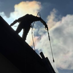 ALIGATOR Alicja Gatlik - Silueta pracownika na dachu z linami, na tle nieba z chmurami. Ujęcie z dołu, podkreślające wysokość i trudność pracy.