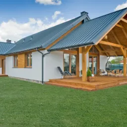 Nowoczesny dom z białymi ścianami i ciemnoszarą metalową konstrukcją dachu, z obszernym drewnianym tarasem i zadaszoną częścią z drewnianymi słupami, krzesłami i wiszącym fotelem.