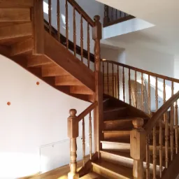 Drewniane schody spiralne z balustradą i toczonymi tralkami, widoczne otwory w ścianie po montażu, na podłodze widoczne refleksy świetlne.