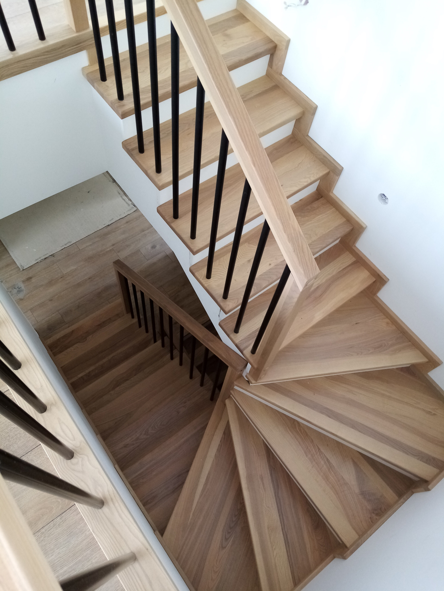 Wysokie ujęcie spiralnych schodów z jasnego drewna z czarnymi balustradami, widoczne fragmenty drewnianej podłogi na dole.