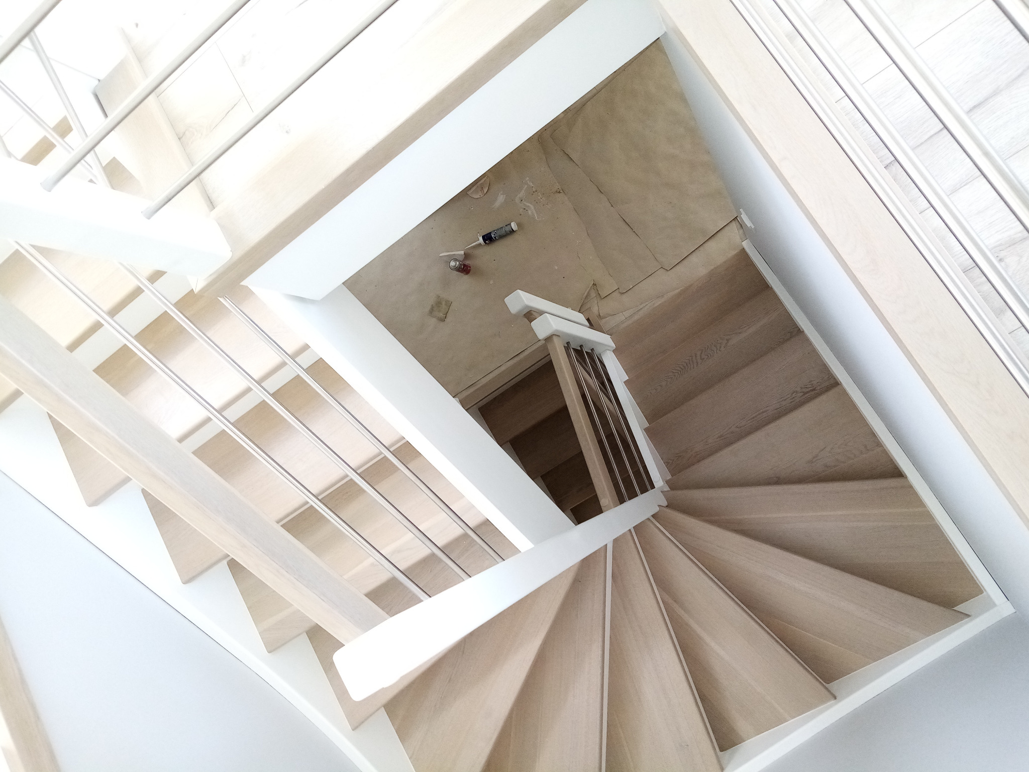 Spiralne schody z jasnego drewna z białymi elementami wykończeniowymi i metalową balustradą, widok z góry na zabezpieczoną podłogę z narzędziami.