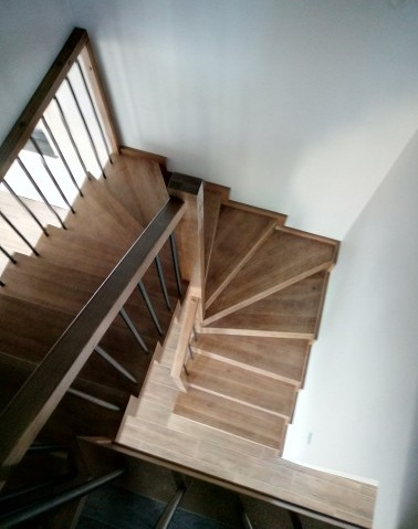 Widok z góry na spiralne schody z jasnego drewna z metalową balustradą, oświetlone naturalnym światłem padającym z góry.