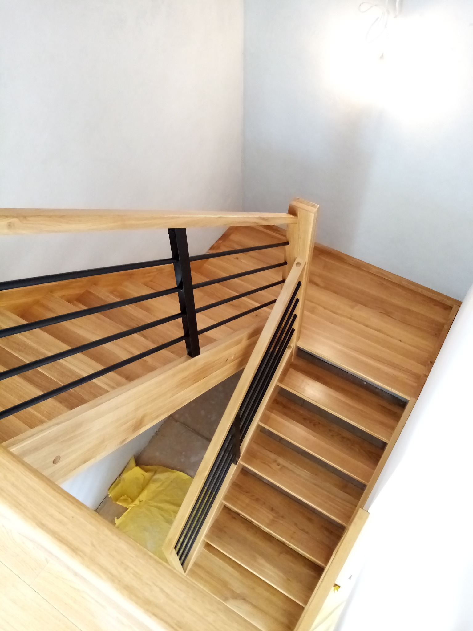 Widok z góry na jasne, drewniane schody z czarnymi, metalowymi balustradami, częściowo zakryte żółtą folią ochronną, w minimalistycznym wnętrzu.