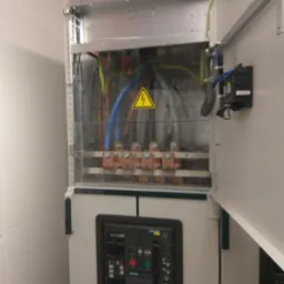 Otwarta szafa elektryczna z widocznymi przewodami, zabezpieczeniami i symbolem ostrzegawczym wysokiego napięcia.