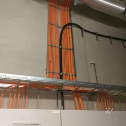 Pomarańczowe kable ułożone w korytkach i przymocowane do ściany, widoczny fragment wentylacji i szafy elektrycznej.