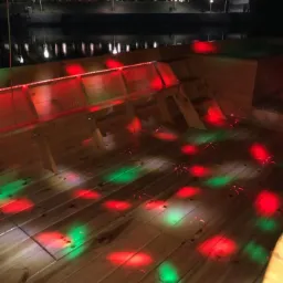 Drewniany podest oświetlony czerwonymi i zielonymi światłami, z widocznymi ławkami i fragmentem zbiornika wodnego w tle.