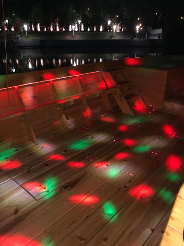 Drewniany podest oświetlony czerwonymi i zielonymi światłami, z widocznymi ławkami i fragmentem zbiornika wodnego w tle.