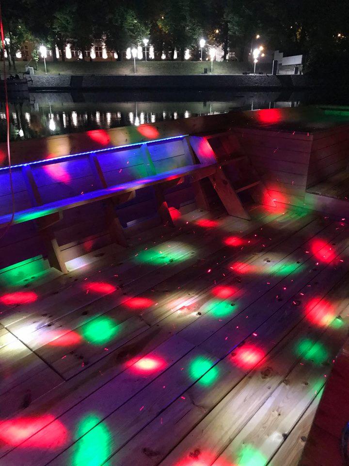 Drewniany podest z ławkami oświetlony czerwonymi i zielonymi światłami dyskotekowymi, w tle oświetlony brzeg rzeki.