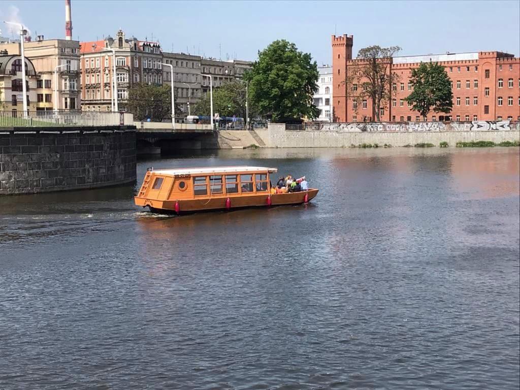 Drewniany statek wycieczkowy płynie rzeką w mieście, z widocznym mostem i budynkami w tle.