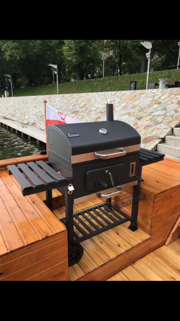 Czarny grill węglowy z bocznymi półkami, umieszczony na drewnianym podeście nad wodą, z polską flagą w tle i kamiennym nabrzeżem.