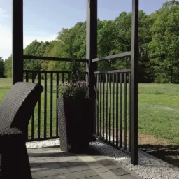 Czarne, pionowe pręty ogrodzenia na tarasie z kostki brukowej, z widokiem na zielony trawnik i drzewa w tle. Donica z fioletowymi kwiatami dodaje koloru.