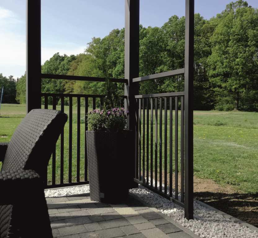 Czarne, pionowe pręty ogrodzenia na tarasie z kostki brukowej, z widokiem na zielony trawnik i drzewa w tle. Donica z fioletowymi kwiatami dodaje koloru.