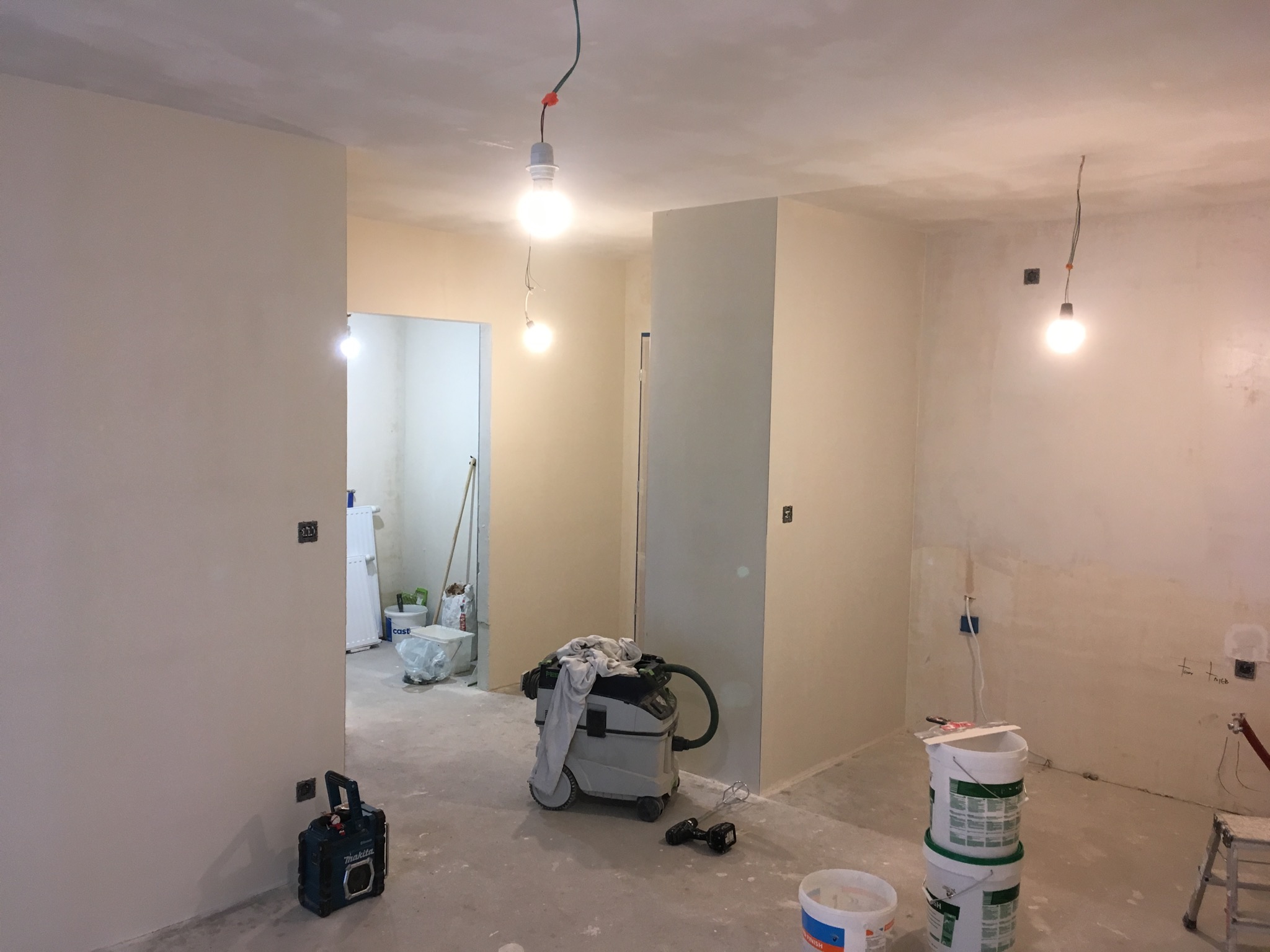 Wnętrze w trakcie prac wykończeniowych z widocznymi śladami szpachlowania na ścianach, wiszącymi żarówkami, odkurzaczem przemysłowym z narzuconą szmatką i stosem wiader po farbie.