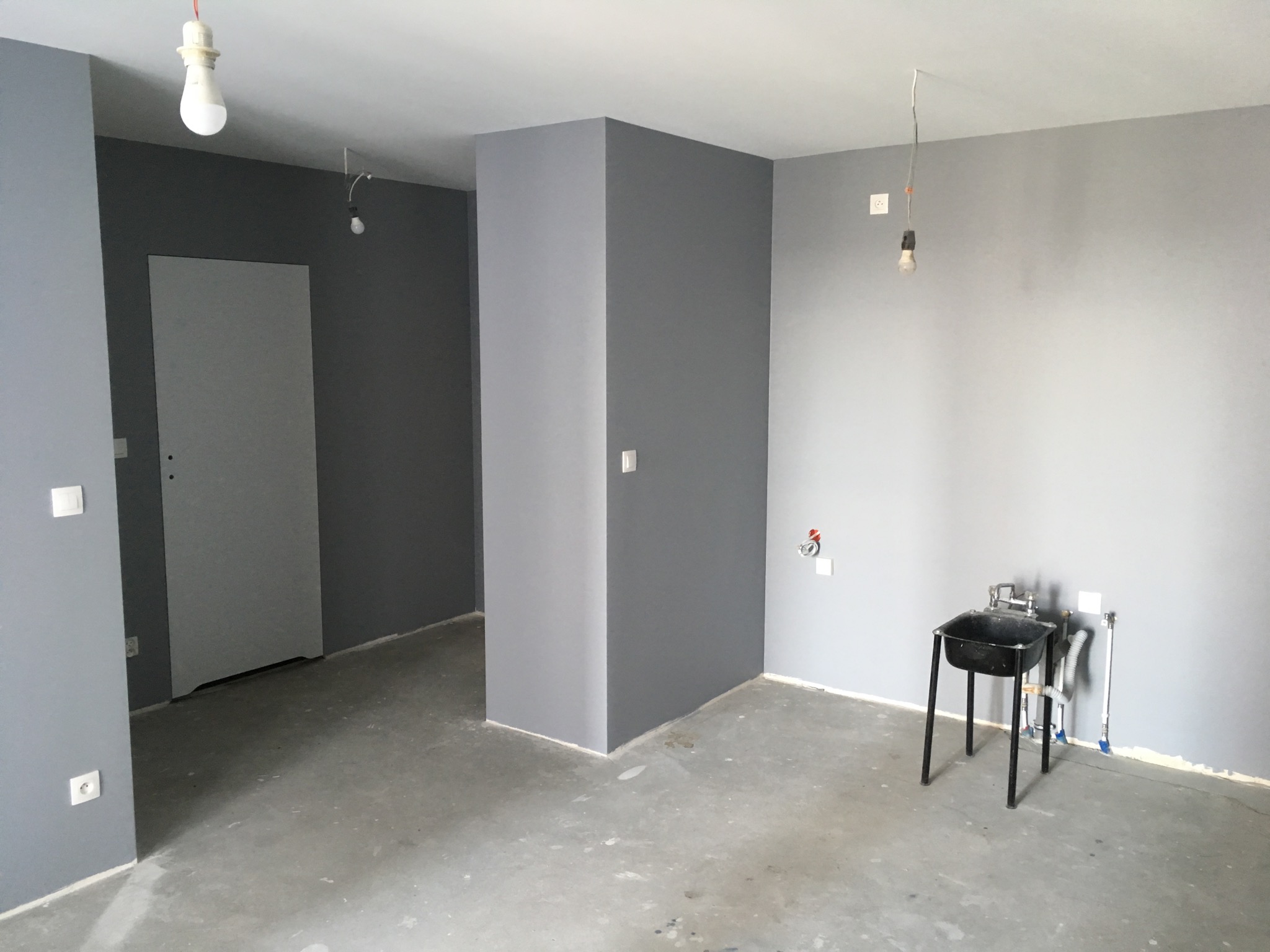 Wnętrze w trakcie prac wykończeniowych, szare ściany, surowa podłoga, prowizoryczne oświetlenie i umywalka na metalowym stelażu.
