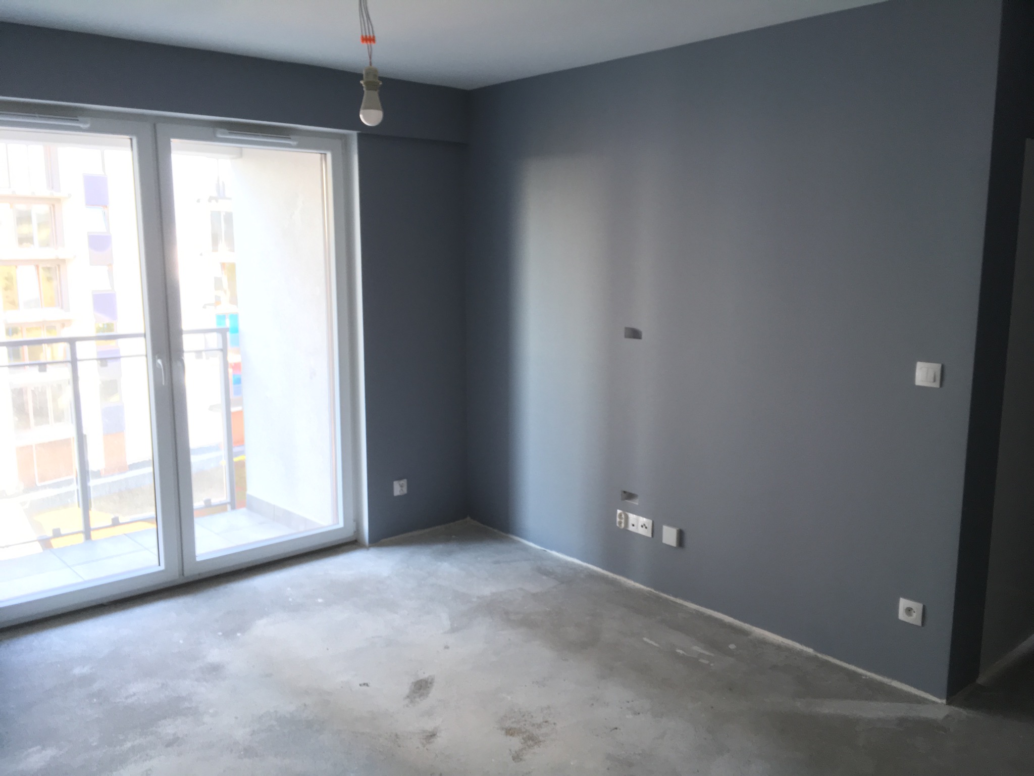 Pomalowane na szaro wnętrze pokoju z dużym oknem balkonowym i surową betonową podłogą przed wykończeniem.