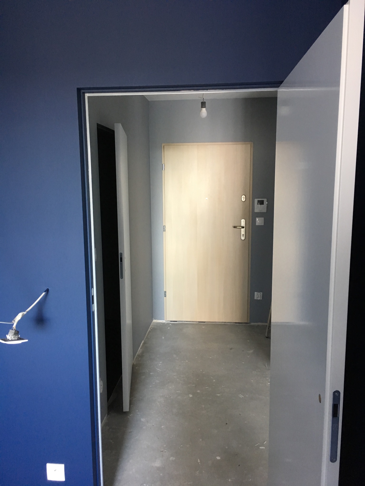 Widok na korytarz z niebieską ścianą i otwartymi białymi drzwiami przesuwnymi, widoczna surowa podłoga i drewniane drzwi wejściowe w tle.