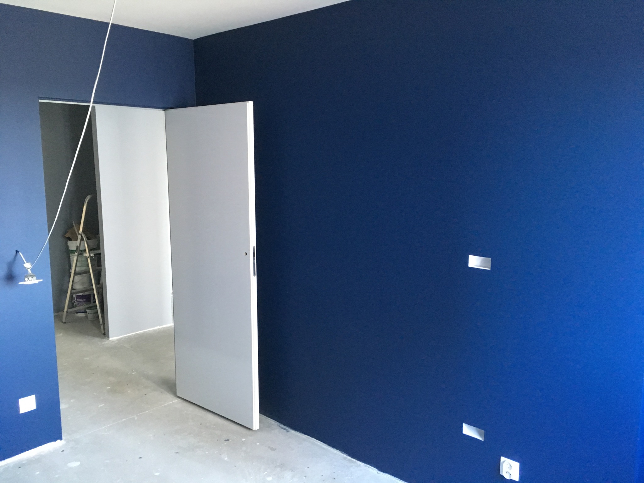 Świeżo pomalowane na granatowo ściany w pokoju z otwartymi białymi drzwiami i widoczną drabiną malarską w tle.