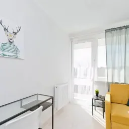 Jasny pokój z biurkiem z szklanym blatem, żółtym fotelem z zieloną poduszką i plakatem z jeleniem w okularach na ścianie.