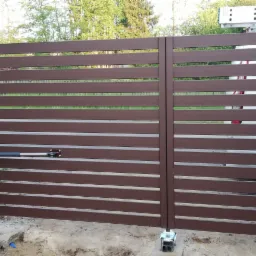 Nowoczesna, brązowa brama przesuwna z automatyką, widoczny siłownik przy słupku i element montażowy przy ziemi, w tle drabina oparta o samochód.