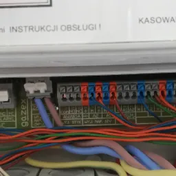 Panel sterowania alarmem domowym z widocznymi przewodami podłączonymi do złączek, etykiety informacyjne i przełącznik w pozycji ON.