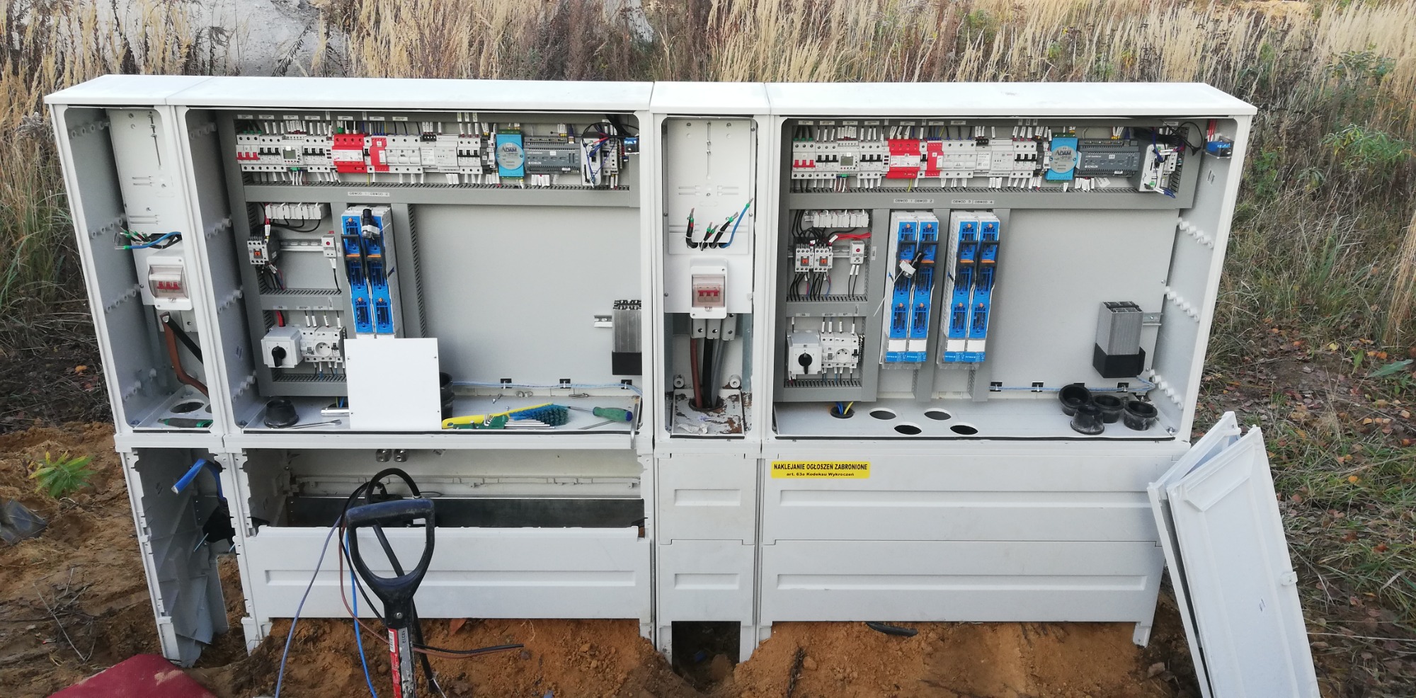 Otwarta, biała szafa elektryczna na zewnątrz, widoczne okablowanie, bezpieczniki i narzędzia wewnątrz, z łopatą i ziemią u podstawy, w tle wysoka trawa.