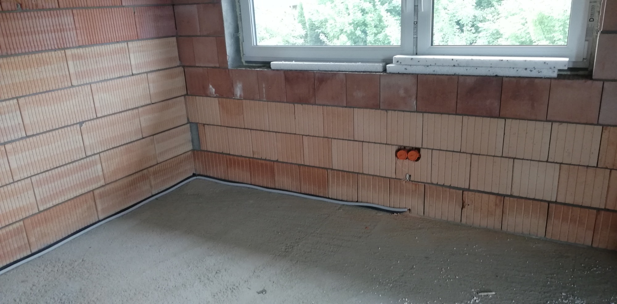 Wnętrze w trakcie budowy z widoczną instalacją elektryczną: pomarańczowe puszki podtynkowe w ścianie z cegły ceramicznej oraz szary przewód elektryczny ułożony wzdłuż ściany na wylewce podłogowej.