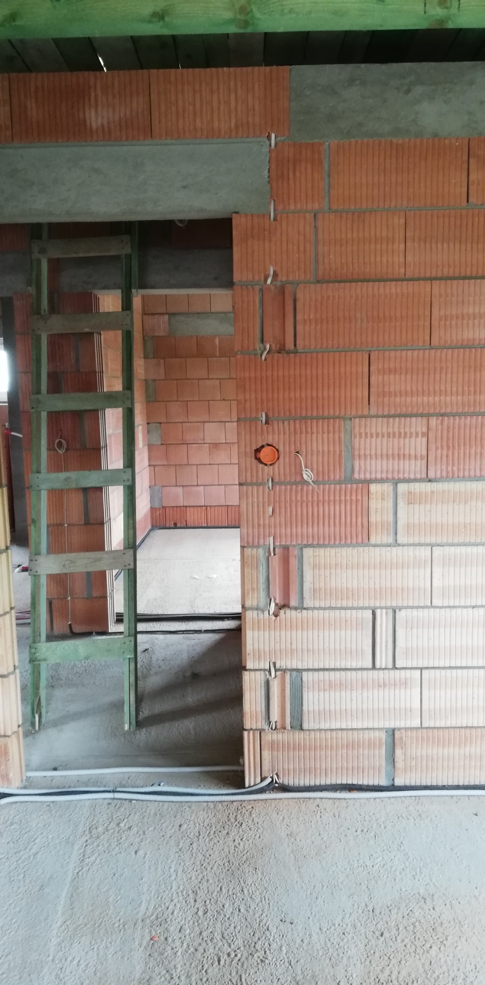 Wewnętrzne ściany z czerwonej cegły dziurawki w trakcie budowy, widoczne przewody elektryczne w peszlach, zielona drabina i otwór na drzwi.
