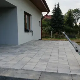 Świeżo ułożona, szara kostka brukowa na posesji domu jednorodzinnego, z żółtą zagęszczarką w tle, widoczne detale fug i krawężników.