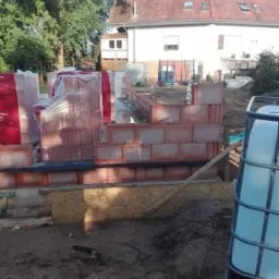 Wznoszenie muru z czerwonej cegły ceramicznej, palety z materiałem w folii, kontener na wodę w tle domu z czerwoną dachówką i zielenią.