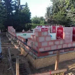 Wznoszenie muru fundamentowego z czerwonej cegły ceramicznej, widoczne materiały budowlane w opakowaniach, szalunki i teren budowy w tle drzewa.