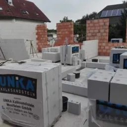 Wznoszenie ścian budynku z bloczków kalksandstein, widoczne palety z materiałem budowlanym marki UNIKA oraz fragmenty murów z czerwonej cegły w tle.